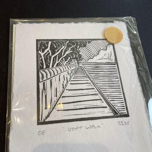 Op de afbeelding: Zwart-wit linoleumsnede getiteld "Goat Walk", met een pad met een leuning, bomen en een verre landschap. Het kunstwerk is gesigneerd en verzegeld met een gouden sticker, gepresenteerd in een transparante beschermhoes.
