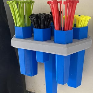 Puede incluir: Un organizador de plástico azul con seis compartimentos para guardar bridas. El organizador tiene una base gris y está montado en una pared. Las bridas son de varios colores, incluyendo verde, negro, rojo y amarillo.