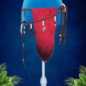 May include: A handcrafted red and brown belt with decorative beads, feathers, and wooden accents. The belt is displayed on a blue mannequin and features the text "Omatikaya". The belt has a braided design and a unique, handcrafted appearance.