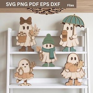 May include: Five layered wooden ghost cutouts displayed on a white shelf. The ghosts feature various accessories including a bow, a basket of apples, a green umbrella, a coffee cup, a green beanie and scarf, a lantern, wheat stalks, and pumpkins.