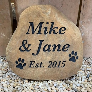 Könnte beinhalten: Ein hellbrauner Stein mit den Namen "Mike & Jane" und dem Jahr "Est. 2015" eingraviert. Zwei schwarze Pfotenabdrücke befinden sich auf beiden Seiten des Textes.