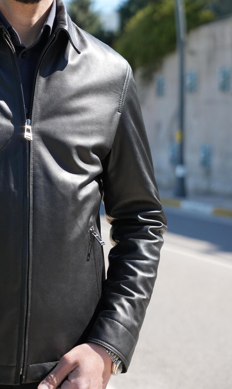 Pu&ograve; includere: Giacca di pelle nera con chiusura a zip e una piccola tasca con zip sulla manica sinistra. La giacca ha un colletto classico e viene indossata sopra una camicia blu scuro. La pelle della giacca appare liscia e lucida.