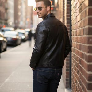 Pu&ograve; includere: Giacca di pelle nera dal design classico. La giacca &egrave; abbinata a jeans blu scuro. La persona indossa occhiali da sole. Lo sfondo comprende un muro di mattoni e una strada cittadina.