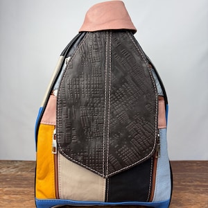 Op de afbeelding: Een patchwork rugzak met gele, blauwe, roze, zwarte en bruine panelen. De flap is van getextureerd donkerbruin leer met witte stiksels. De bovenkant is lichtroze.