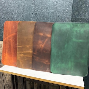 Può includere: Quattro tappetini da scrivania in pelle di diversi colori: arancione, marrone, marrone scuro e verde. I tappetini sono allineati su una superficie bianca.