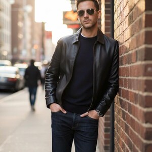 Pu&ograve; includere: Giacca di pelle nera con collo e chiusura a zip. La giacca &egrave; abbinata a un maglione blu scuro e jeans scuri. La persona indossa occhiali da sole da aviatore e un orologio. Lo sfondo comprende un muro di mattoni e una strada cittadina.