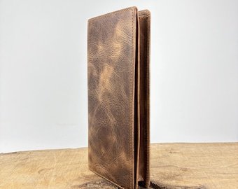 Op de afbeelding: Een verticale, bruine leren portemonnee met een distressed textuur. De portemonnee staat rechtop en toont het gevouwen ontwerp en zichtbare stiksels. Het leer heeft een versleten uiterlijk, wat duurzaamheid en gebruik suggereert.