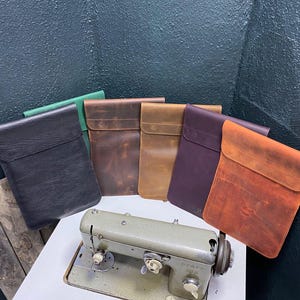 Può includere: Sei custodie per laptop in pelle di diversi colori, tra cui nero, marrone, verde e viola, sono disposte su un tavolo davanti a una macchina da cucire. Le custodie sono realizzate in pelle di alta qualità e hanno un design semplice ed elegante.