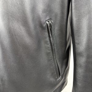 Pu&ograve; includere: Primo piano di una giacca di pelle nera con tasca con cerniera. La giacca ha una finitura liscia e opaca e un design classico. La cerniera della tasca &egrave; argentata.