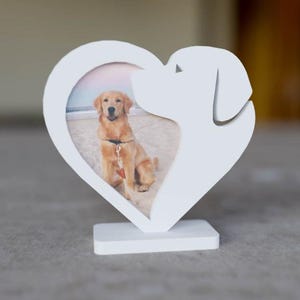 Puede incluir: Un marco de fotos blanco en forma de corazón con una silueta de perro en el lado derecho. El marco contiene una foto de un golden retriever. El marco se apoya en una base rectangular.
