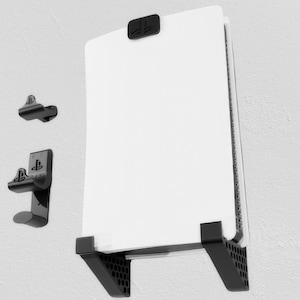 May include: A white PlayStation 5 console mounted on a black wall mount with two black controllers mounted on a separate wall mount.
