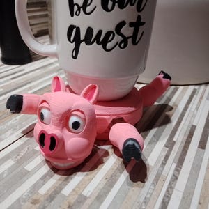 Puede incluir: Un soporte para tazas con forma de cerdo rosa que sostiene una taza blanca con el texto "be our guest" en negro. El cerdo tiene pezuñas negras y una cara detallada. Un accesorio de cocina caprichoso.