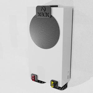 May include: White Xbox Series S console mounted on a wall. The console has a black top piece with the Xbox logo and a circular speaker grill. Red and yellow accents are visible on the bottom.