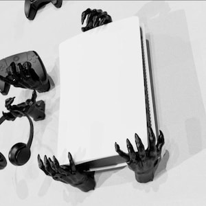 May include: Black wall-mounted gaming console holder with matching controller and headset holders. The console is held by two black claw-like hands. The controller and headset are held by similar hands.