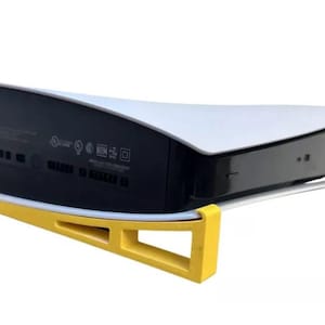 Puede incluir: Un soporte de plástico amarillo para una consola PlayStation 5 blanca. El soporte está diseñado para sostener la consola verticalmente y tiene una forma curva para adaptarse al diseño de la consola.