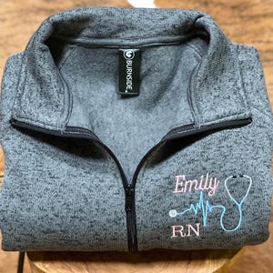 May include: Grey fleece zip-up sweatshirt with a pink embroidered design that says "Emily RN" with a stethoscope and a heart beat line.