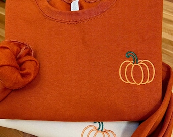 May include: An orange sweatshirt featuring an embroidered pumpkin design with an orange outline and a green stem. Another sweatshirt with a similar pumpkin design is visible underneath. The sweatshirts are folded on a wooden surface.