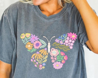 May include: A dark grey t-shirt featuring a colourful floral butterfly design. The butterfly is made up of pink, purple, yellow, orange, and green flowers.