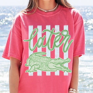 Puede incluir: Camiseta de color coral con un gr&aacute;fico de un caim&aacute;n verde y la palabra "later" en una fuente cursiva. El dise&ntilde;o incluye rayas blancas verticales. La camiseta tiene mangas cortas y un corte holgado.