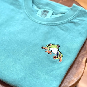 May include: A teal-colored t-shirt with a small embroidered frog on the front. The frog has a green head, white belly, and orange feet. The shirt is folded and resting on a wooden surface. The brand name is visible on the tag.