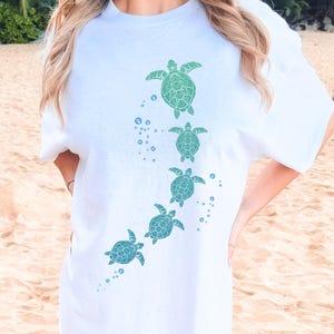 May include: White t-shirt featuring a design of five sea turtles in shades of green and blue, ascending diagonally. Bubbles accent the turtles' path. The shirt is a casual, comfortable style.