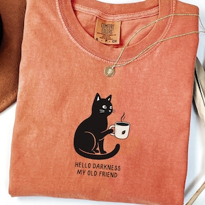 May include: Coral-colored Comfort Colors t-shirt featuring a black cat holding a coffee cup. The shirt has the phrase "HELLO DARKNESS MY OLD FRIEND" printed below the cat illustration. A gold necklace is draped across the shirt.