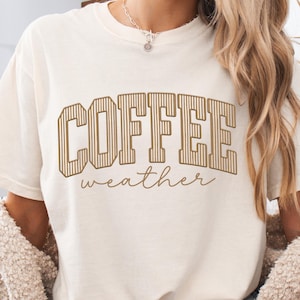 May include: A cream-coloured short-sleeve t-shirt featuring the text COFFEE in large, brown, vertically striped block letters, with the word weather written below in a smaller, brown, cursive script font. The shirt has a crew neckline.