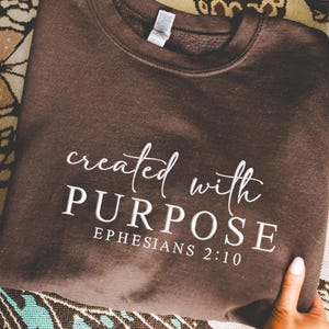 May include: A dark brown sweatshirt with the words "created with PURPOSE" and "EPHESIANS 2:10" printed in white script. The sweatshirt is displayed on a patterned surface.
