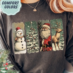 May include: Dark grey sweatshirt with a Christmas-themed graphic. The graphic features a snowman, Christmas tree, Santa Claus, and a reindeer. The text "COMFORT COLORS" is in a light blue circle in the upper left corner.