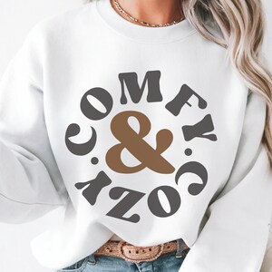 May include: White sweatshirt featuring the text "COMFY & COZY" in a circular arrangement. The "&" is brown, while the other letters are dark grey. The sweatshirt is a relaxed, casual style.