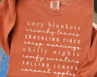 May include: Rust-colored long-sleeve sweatshirt with white text listing fall-themed phrases like "cozy blankets" and "falling leaves." The sweatshirt has a crew neck and a Comfort Colors tag. The sleeve is partially visible.