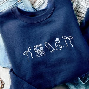 May include: Navy blue sweatshirt with white line art of a bow, computer, spray bottle, phone, and bow. The sweatshirt is folded on top of blue jeans and a white knitted blanket.