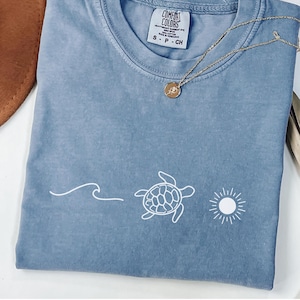 May include: A light blue t-shirt with a white graphic of a turtle swimming in the ocean with a sun above it. The shirt has a tag that says "Comfort Colors" and "S - P - CH".
