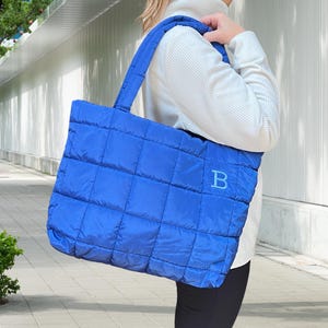 May include: A vibrant blue quilted tote bag with a stitched grid pattern. The bag features a matching blue handle and a large letter "B" embroidered on the front. The bag is made of a soft, padded material.