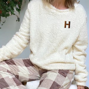 May include: Cream-colored, fuzzy sweatshirt with a brown letter "H" on the chest. The sweatshirt is paired with brown and cream plaid pajama pants. The outfit appears soft and comfortable, suitable for lounging.