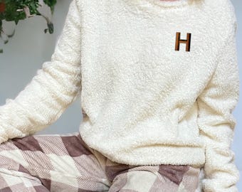 May include: A cream-coloured, long-sleeved, fuzzy fleece pullover jumper featuring a crew neckline and a small, block-lettered H monogram on the left chest. The jumper is paired with brown and cream plaid patterned lounge trousers.