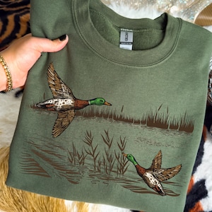 K&ouml;nnte beinhalten: Ein olivgr&uuml;nes Sweatshirt mit einer detaillierten Illustration von zwei Enten im Flug &uuml;ber Wasser und Schilf. Die Enten haben braune, wei&szlig;e und gr&uuml;ne Markierungen. Ein bequemes, l&auml;ssiges Kleidungsst&uuml;ck.