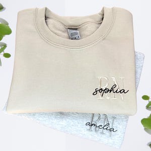 Puede incluir: Dos sudaderas beige y grises con las letras "RN" bordadas en la parte delantera. La sudadera superior tiene el nombre "sophia" bordado debajo de las letras. La sudadera inferior tiene el nombre "amelia" bordado debajo de las letras.