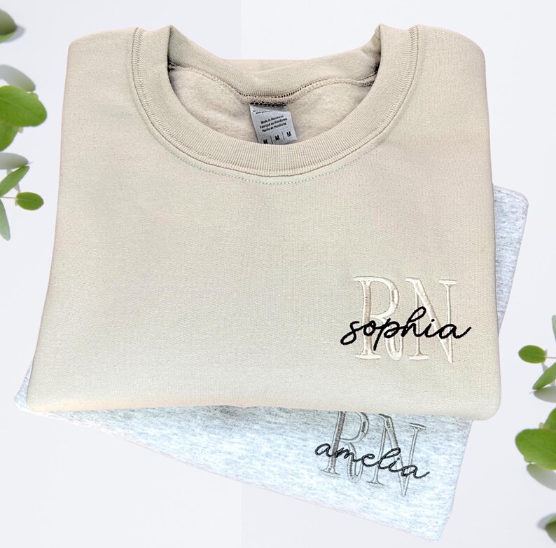 Puede incluir: Dos sudaderas beige y grises con las letras "RN" bordadas en la parte delantera. La sudadera superior tiene el nombre "sophia" bordado debajo de las letras. La sudadera inferior tiene el nombre "amelia" bordado debajo de las letras.