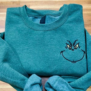 Puede incluir: Sudadera verde azulado con un rostro del Grinch bordado. La cara del Grinch est&aacute; delineada en negro con ojos amarillos. La sudadera tiene cuello redondo y mangas largas. La tela parece ser un material suave y c&oacute;modo.