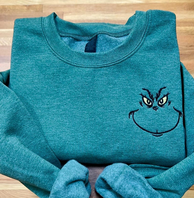 Puede incluir: Sudadera verde azulado con un rostro del Grinch bordado. La cara del Grinch est&aacute; delineada en negro con ojos amarillos. La sudadera tiene cuello redondo y mangas largas. La tela parece ser un material suave y c&oacute;modo.
