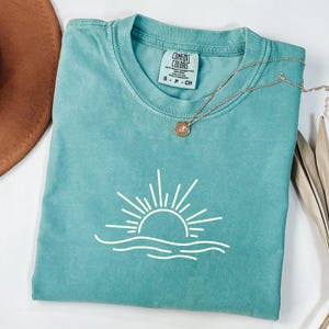 May include: A teal Comfort Colors t-shirt with a white graphic of a sun over water. The shirt has a crew neck and short sleeves. A gold necklace with a small pendant is draped over the shirt.