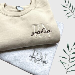 Puede incluir: Dos sudaderas con texto bordado. La sudadera de arriba es beige con el texto "RN sophia" en blanco. La sudadera de abajo es gris con el texto "RN amelia" en negro.