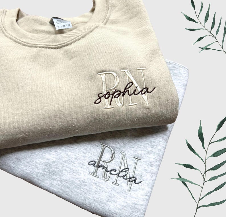 Puede incluir: Dos sudaderas con texto bordado. La sudadera de arriba es beige con el texto "RN sophia" en blanco. La sudadera de abajo es gris con el texto "RN amelia" en negro.