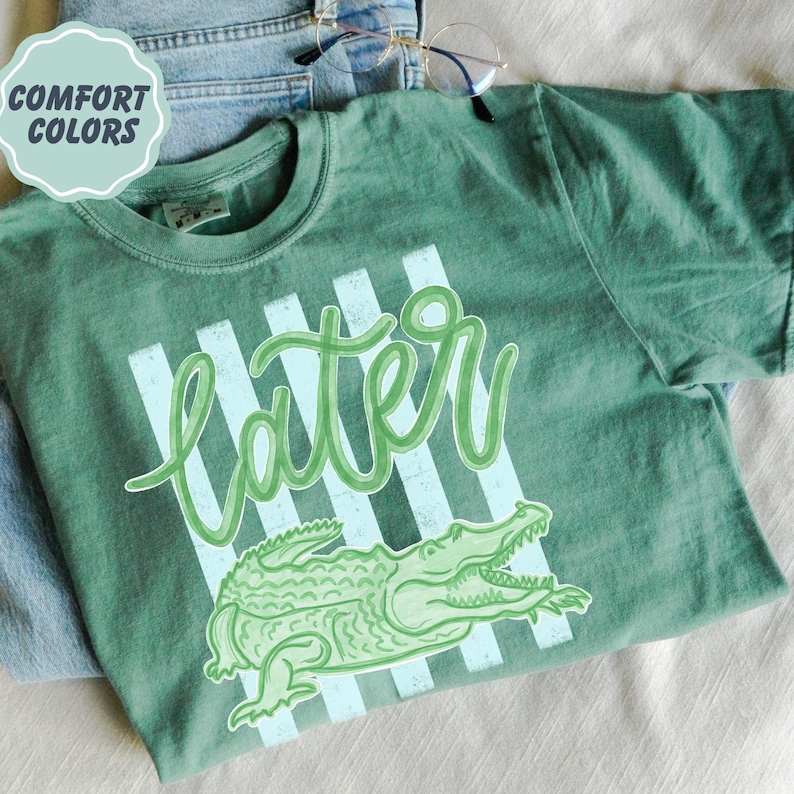 Puede incluir: Camiseta verde con la palabra "Later" en fuente cursiva y un dibujo de un cocodrilo. La camiseta tiene cuello redondo y mangas cortas. Las palabras "Comfort Colors" est&aacute;n en un c&iacute;rculo verde claro en la esquina superior izquierda.