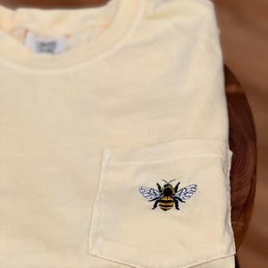 May include: A pale yellow short-sleeved t-shirt featuring a single chest pocket. The pocket is adorned with a detailed embroidered graphic of a bumblebee with black and yellow stripes and white wings. A white fabric label is visible at the neckline.