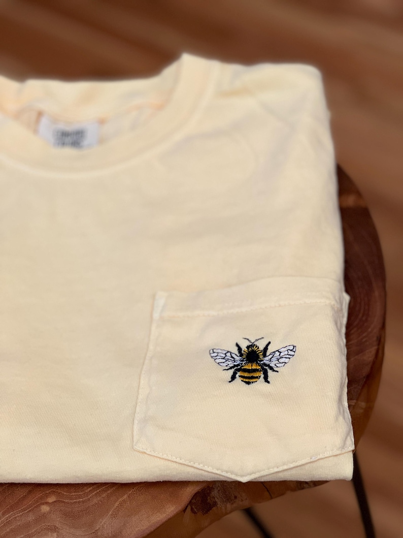 May include: A pale yellow short-sleeved t-shirt featuring a single chest pocket. The pocket is adorned with a detailed embroidered graphic of a bumblebee with black and yellow stripes and white wings. A white fabric label is visible at the neckline.