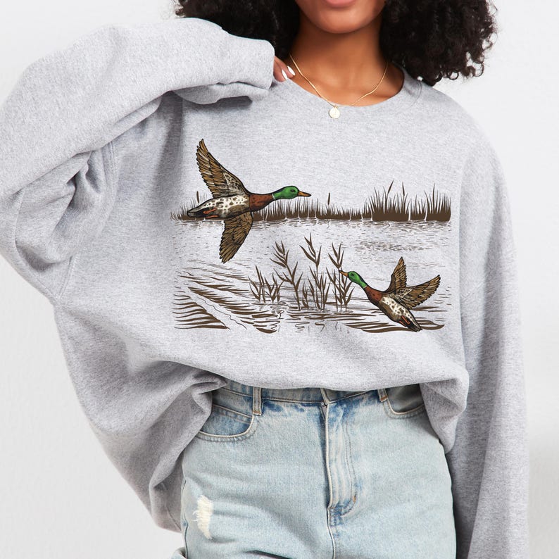 K&ouml;nnte beinhalten: Hellgraues Sweatshirt mit detaillierter Illustration von zwei Enten im Flug &uuml;ber Wasser und Schilf. Die Enten sind braun, gr&uuml;n und wei&szlig;. Das Sweatshirt ist ein l&auml;ssiger Stil.