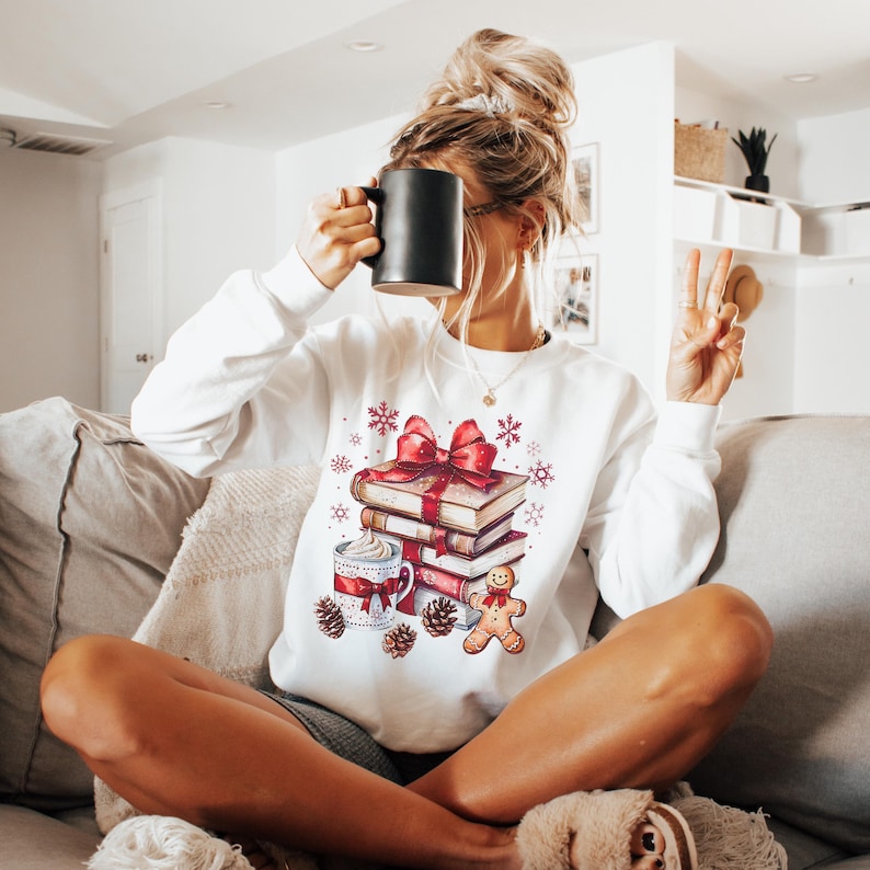 May include: White long-sleeve sweatshirt with a festive design. The graphic features a stack of books tied with a red bow, a mug of hot cocoa, a gingerbread man, pine cones, and snowflakes. A black mug is held up.