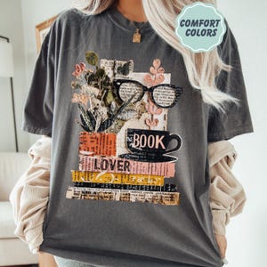 May include: Dark gray t-shirt with a graphic design featuring a stack of books, a coffee cup, and eyeglasses. The word "BOOK" is printed on the cup, and "LOVER" is on the books. The shirt has the words "COMFORT COLORS" in a light blue circle.